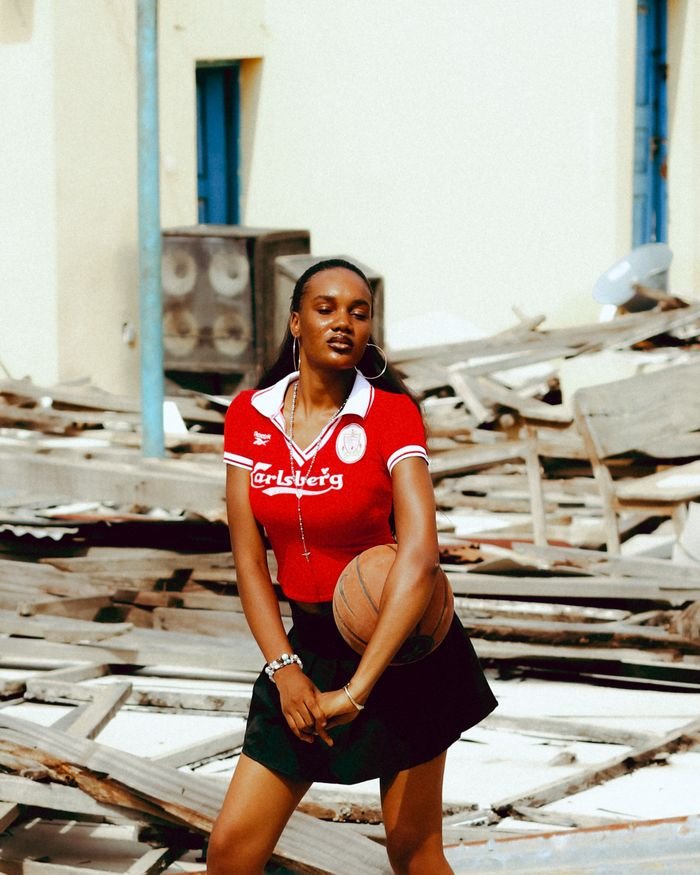 Young woman with a basketball poses confidently outdoors amidst urban debris, wearing a red sports shirt and hoop earrings.